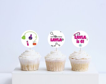 May include: Three white cupcakes with pink and white frosting. Each cupcake has a round, white paper topper with a science theme. The toppers feature a beaker, test tube, and atom, along with the text "Happy Birthday Layla" and "Layla is 6!"