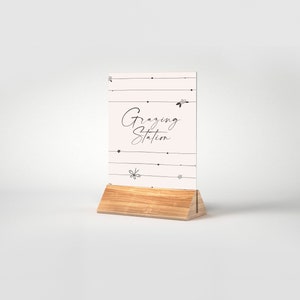 May include: A white sign with black text and floral accents that reads "Grazing Station" on a wooden stand.