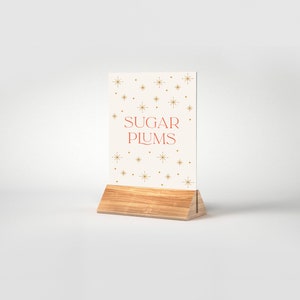 May include: A white sign with gold stars and the text "SUGAR PLUMS" in red letters. The sign is on a wooden stand.