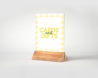 May include: A yellow and white gingham sign with the text "CARDS and GIFTS" in yellow letters. The sign is on a wooden stand.