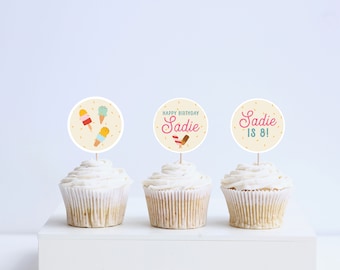 Könnte beinhalten: Drei Cupcakes mit weißem Zuckerguss und dekorativen Toppern. Ein Topper zeigt Eis-Illustrationen, während die anderen "Happy Birthday Sadie" und "Sadie is 8!" in Rosa und Türkis zeigen. Die Cupcakes stehen auf einer weißen Oberfläche.