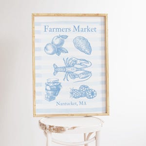 Peut inclure: Une affiche encadrée avec un fond à rayures bleu clair et blanc. L'affiche présente des illustrations de fruits, d'un homard et d'autres articles, avec le texte "Farmers Market" et "Nantucket, MA" en bleu.