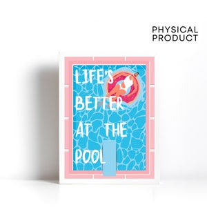 May include: A pink framed print with a blue and white pool scene. The print says "Life's Better At The Pool" with a person floating on a pink donut.