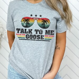 May include: A heather grey t-shirt with a retro-style graphic of sunglasses with palm trees and the text "TALK TO ME GOOSE".