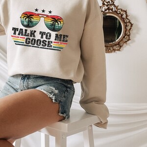 以下が含まれることがあります： パームツリーが描かれたサングラスと「Talk to me Goose」というテキストがレトロなスタイルでプリントされたベージュのトレーナーです。