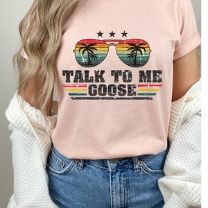 May include: A pink t-shirt featuring a retro-style graphic of sunglasses with palm trees reflected in the lenses. The text "TALK TO ME GOOSE" is printed below the sunglasses.