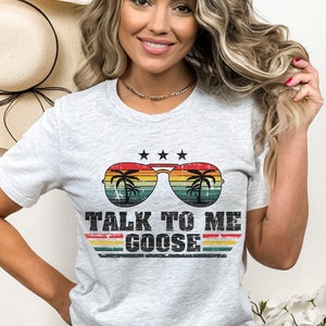 May include: A grey t-shirt featuring a vintage-style graphic of sunglasses with palm trees and the text "TALK TO ME GOOSE" in a distressed font.
