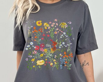 以下が含まれることがあります： 鮮やかな花柄が特徴のアスファルトカラーのTシャツ。黄色、赤、紫の花など、様々な色のワイルドフラワーが描かれています。このシャツは半袖でゆったりとしたフィット感があり、カジュアルウェアに最適です。
