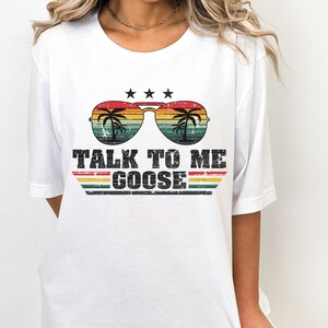 May include: White t-shirt featuring a retro-style graphic of sunglasses with palm trees reflected in the lenses. The text "TALK TO ME GOOSE" is printed below the sunglasses.