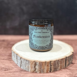 Op de afbeelding: Een bruine glazen pot met een zwart deksel, met het label "Frankenstein" van Main Street Mercantile. Het turquoise label heeft sierlijke ontwerpen en geurnoten. De pot staat op een natuurlijke houtsnipper, tegen een donkere achtergrond.