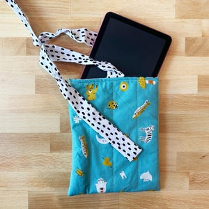 May include: A teal quilted tablet sleeve with a black and white polka dot strap. The sleeve features a repeating pattern of yellow fish, zebras, and other whimsical illustrations. A black tablet is inside the sleeve.