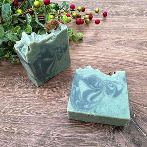 May include: Two bars of handmade soap with a green and gray marbled design. The soap is sitting on a wooden surface.
