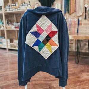 Puede incluir: Una sudadera con capucha azul marino con un diseño de bloque de colcha colorido en la espalda. El bloque de colcha presenta un patrón de estrella con varios colores, incluyendo rosa, rojo, azul y amarillo. La sudadera se muestra en una percha blanca.