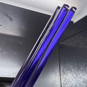 May include: Four long, thin, dark blue glass rods are stacked together. The rods are smooth and shiny, and they are illuminated by a bright light source.