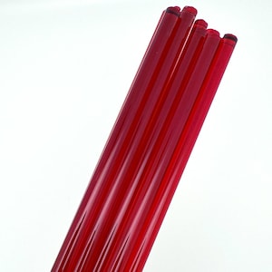 May include: A cluster of six long, cylindrical, red glass rods. Each rod has a ribbed design along its length. The rods are arranged diagonally against a white background, showcasing their vibrant color and texture.
