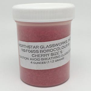 May include: A small, clear plastic jar filled with red glass frit. The jar has a white label with the text "NORTHSTAR GLASSWORKS, INC NS-F065S BOROCOLOUR® CAUTION: AVOID BREATHING GLASS DUST CHERRY SIZE S 4 OUNCES (112 GRAMS)"