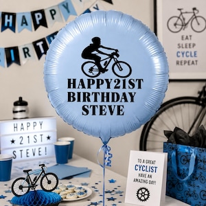 May include: A round, light blue foil balloon featuring a black silhouette of a cyclist on a mountain bike and the black text "HAPPY 21ST BIRTHDAY STEVE". The balloon has a metallic finish and is attached to a blue ribbon at the bottom.