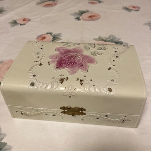 May include: A white jewelry box with a floral design. The box has a pink rose with gold accents and a gold latch.