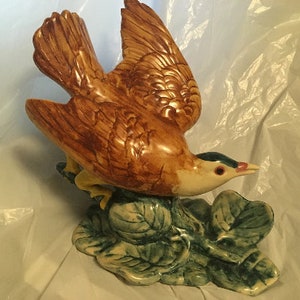 Puede incluir: Una figurita de pájaro de cerámica con plumas marrones y amarillas, posada sobre una base de hojas verdes. El pájaro tiene un pico largo con una punta azul y roja.