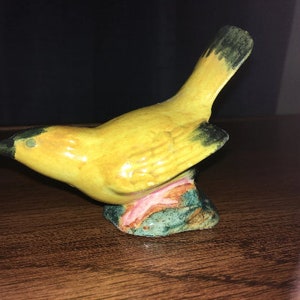 Könnte beinhalten: Eine kleine Keramik-Vogelfigur mit gelben und schwarzen Details. Der Vogel sitzt auf einem Sockel, der mit Grün, Rosa und Orange bemalt ist.