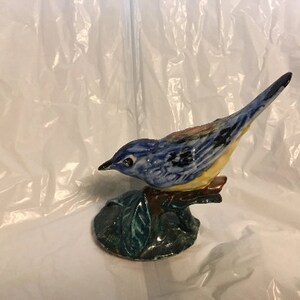 May include: A blue and yellow ceramic bird figurine perched on a green leafy base. The bird has black markings on its wings and a detailed beak.