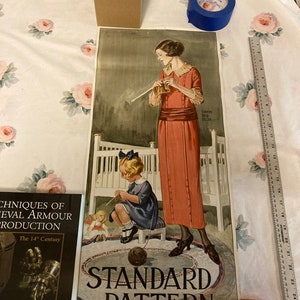 May include: A vintage poster advertising Standard Pattern dress and apron designs. The poster features a woman and a young girl knitting, with the text "Standard Pattern" and the dress and apron numbers, "Dress 1549" and "Apron 9397".