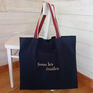 Peut inclure: Un sac fourre-tout bleu marine avec le texte "Sous les étoiles" en écriture cursive. Le sac a des anses rayées rouges, blanches et dorées. Le sac est posé sur une chaise blanche.