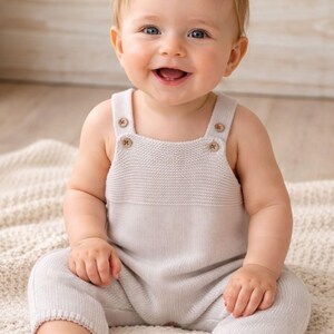Op de afbeelding: Een lachende baby in een lichtgrijze gebreide romper met houten knoopjes. De romper heeft een vierkante halslijn en korte pijpen. De baby zit op een crèmekleurige deken.