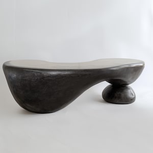 May include: A dark grey, organically shaped bench with a smooth, curved surface. The bench is supported by a rounded base and a single, spherical leg. The design is modern and sculptural, suitable for interior decor.