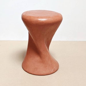 May include: A terracotta-colored ceramic stool with a twisted, organic shape. The stool has a smooth, rounded top and a wide base.