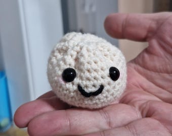 May include: A crocheted white stuffed toy shaped like a bun with a smiling face. The toy has black eyes and a black mouth.