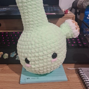 May include: A handmade, plush, light green bong with a cute face, black eyes, and pink cheeks. The top and mouthpiece are white. The bong is on a blue sticky note on a wooden surface.