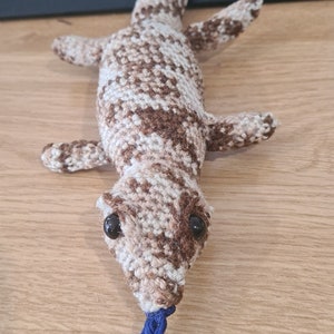 Puede incluir: Un juguete de gecko de crochet marrón y blanco con una cola azul. El gecko tiene ojos negros y un cuerpo con estampado marrón y blanco.