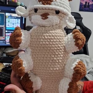 May include: A handmade crochet cow plush toy. The toy is mainly beige with white and brown details on the head, ears, and hooves. The cow has a smiling face and wears a white and brown hat. The toy is made with soft yarn.