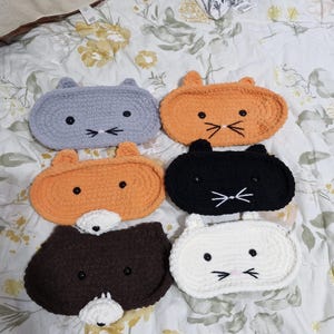 May include: Five crocheted cat-shaped eye masks in different colours: grey, orange, black, brown, and white. Each mask has embroidered eyes, a nose, and whiskers.