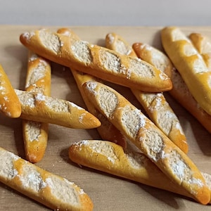May include: A selection of golden-brown baguettes, lightly dusted with white flour, are displayed on a light brown surface. The bread has a textured crust with diagonal cuts. The image showcases a collection of freshly baked bread.