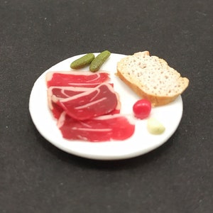 Op de afbeelding: Een wit bord met een portie gesneden vlees, een snee brood, twee groene augurkschijfjes en een rode cherrytomaat.