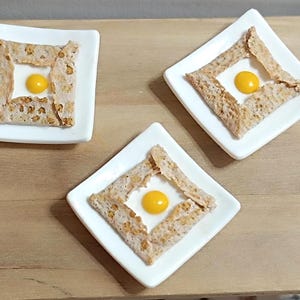 Puede incluir: Tres platos cuadrados blancos sostienen cada uno un crepe salado, doblado para enmarcar una yema de huevo amarilla brillante. Los crepes tienen un color marrón claro moteado, y los platos están sobre una superficie de madera marrón claro.
