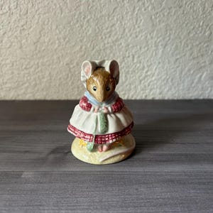 May include: A ceramic figurine of a mouse wearing a white dress with red trim and a matching bonnet. The mouse has brown fur and is standing on a yellow base. The figurine is a decorative item.