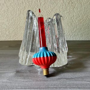 May include: A clear, textured glass candle holder with a vertical, wavy design. Positioned in front is a decorative light bulb featuring a tall, thin red glass tube, a blue and red ribbed base, and a standard metal screw-in electrical connector.