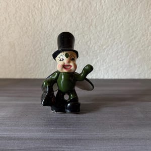 May include: A vintage figurine of a smiling, green-bodied character wearing a black top hat and shoes. The figurine has a white face with red cheeks and a black bag. The figure is standing on a gray surface.