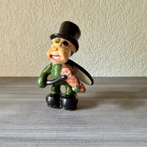 May include: A whimsical ceramic figurine of a green insect-like character wearing a black top hat. The figure has a smiling face, black boots, and holds a brown object. The wings are black and the figurine is on a gray surface.