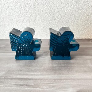 May include: Two teal-colored angel candle holders. Each angel has detailed wings and a small circular indentation for a candle. The candle holders are made of a glossy material and sit on a wooden surface.