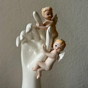 May include: A white ceramic hand sculpture holds two cherubic figurines. The cherubs have blonde hair, small wings, and are posed as if climbing the fingers. The sculpture has a matte finish and is set against a neutral background.