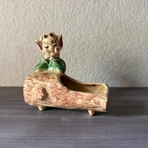 May include: A vintage ceramic planter shaped like a log, with a whimsical elf figure perched on top. The elf has a cream-colored face, pink ears, and is wearing a green top. The log planter is beige and brown.