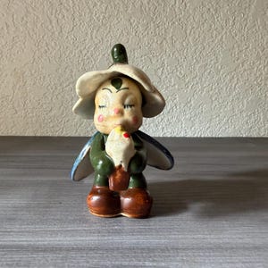 May include: A ceramic figurine of a whimsical fairy-like creature. It has a white flower hat, green body, blue wings, and brown boots. The figurine is holding a white object with a red accent.