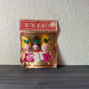 May include: A package of three felt Christmas ornaments featuring stylized doll figures with yellow and brown hair, pink faces, and colorful dresses. The packaging is labeled CHRISTMAS DECORATIONS in red text on a gold background with a clear plastic window.