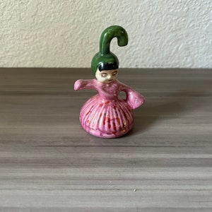 May include: A small ceramic figurine of a person in a pink dress with a green hat. The figurine has a detailed face and arms outstretched. The dress has vertical lines and red accents. The hat is a curved shape.