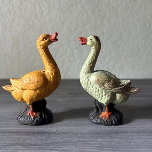 May include: Two goose figurines. One is yellow with a red beak and feet, the other is off-white with a red beak and feet. Both are perched on a black base. The geese face each other, creating a charming display.