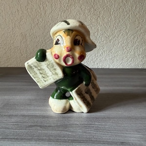 May include: A vintage ceramic figurine of a newsboy, wearing a white cap and green jacket. The figurine has a surprised expression with rosy cheeks and is holding newspapers. The base of the figurine is off-white.